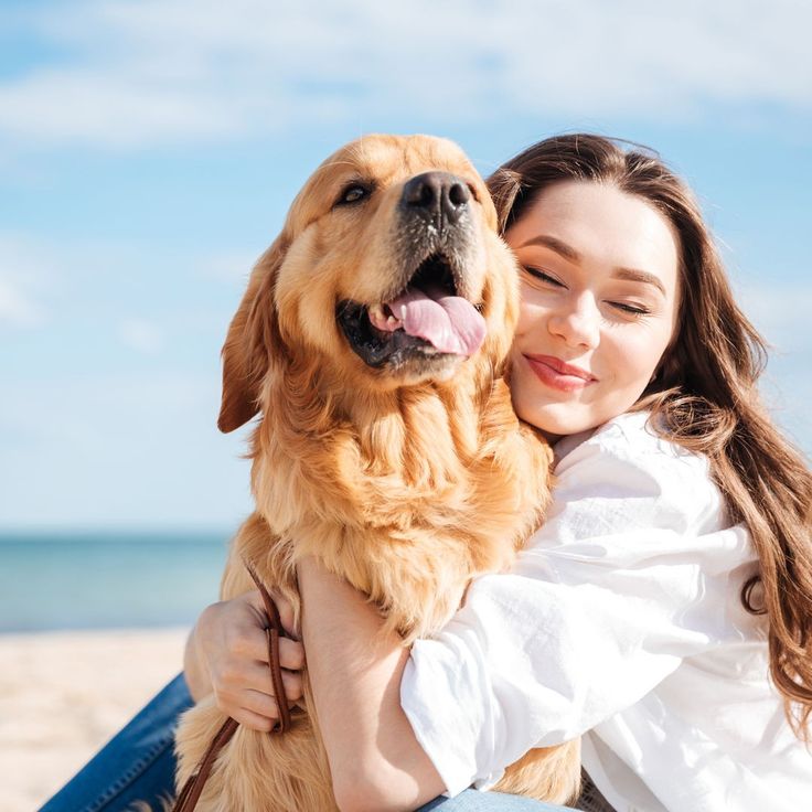 A smiling young woman with long brown hair hugging a happy golden retriever dog on a sunny beach with a blue sky background.