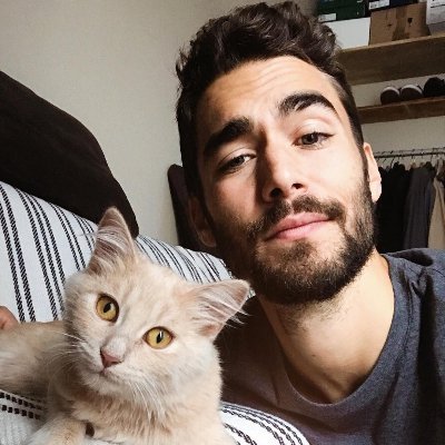 A smiling, bearded young man with dark hair lying down next to a light-colored, fluffy cat on a striped pillow.